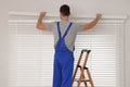 Worker in uniform installing horizontal window blinds on stepladder indoors, back view