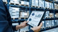 A worker in a warehouse stands in an aisle and looks at a tablet that displays a data chart Royalty Free Stock Photo