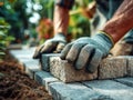 Worker wearing protective gloves carefully placing stone bricks to construct a garden pathway surrounded by soil and greenery in a Royalty Free Stock Photo