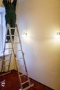 Worker in workwear is installing a chandelier and connects it to electricity on the ceiling from ladder in apartment is Royalty Free Stock Photo