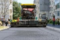 Workers operating asphalt paver machine during road repairs in front of a multi-storey residential building on a sunny summer day