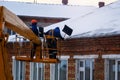 Workers in overalls and orange helmets on the crane basket remove icicles from roof of the house on a winter day - cleaning the