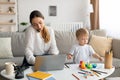 Working with baby. Busy young mother talking on cellphone and typing on laptop while her toddler son playing nearby Royalty Free Stock Photo