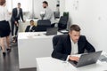 Working day in the office, young man sits at a table and works on laptop in the office, his colleages stands beside him