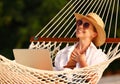 Young happy woman wearing in straw hat and sunglasses enjoying freelance work on laptop while lying in the hammock on the beach Royalty Free Stock Photo