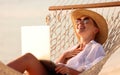 Young happy woman wearing in straw hat and sunglasses enjoying freelance work on laptop while lying in the hammock on the beach Royalty Free Stock Photo