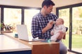 Working Father Using Laptop At Home Whilst Feeding Baby Son On Knee With Bottle Royalty Free Stock Photo