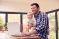 Working Father Using Laptop At Home Whilst Holding Smiling Baby Son On Knee Royalty Free Stock Photo