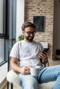 Working at home. Cheerful young handsome man drinking coffee talking on the mobile phone while sitting in big comfortable chair at Royalty Free Stock Photo