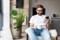 Working at home. Cheerful young handsome man drinking coffee talking on the mobile phone while sitting in big comfortable chair at Royalty Free Stock Photo