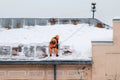 A working man in bright overalls with a safety belt with a shovel clears snow from the roof of an old building