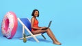 Working online from tropical paradise. Black woman in bikini sitting in lounge chair, using laptop on blue background Royalty Free Stock Photo