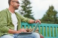 Working in the park. Young man wearing eyeglasses sitting on the bench outdoors and using laptop Royalty Free Stock Photo