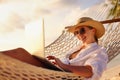 Working at sunset. Side view of young happy female using laptop while lying in the hammock on the beach Royalty Free Stock Photo