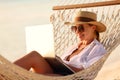 Working at sunset. Side view of young happy female using laptop while lying in the hammock on the beach Royalty Free Stock Photo