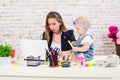 Cheerful young beautiful businesswoman looking at laptop while sitting at her working place with her little daughter Royalty Free Stock Photo