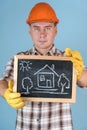 A workman in an orange helmet holds a chalk board with a comic drawing of a house at arm`s length