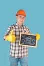 A workman in an orange helmet shows a thumbs up holding a chalk board with a comic drawing of a house in his hand