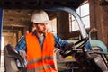 Workman wears protective helmet, vest and gloves. Worker standing near forklift in warehouse.