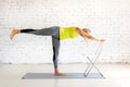 Workout with props. Fit caucasian woman practice yoga balance with one leg up and a chair on a mat in loft white studio