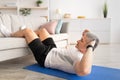 Workout for vitality. Athletic mature man doing exercises for abs, putting feet on sofa, exercising on mat at home