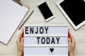 Workplace with tablet, smartphone, notepad and `Enjoy today` word on lightbox over white wooden background, top view. Female han Royalty Free Stock Photo