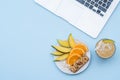 Workspace with laptop and with a healthy breakfast: mango, orange and muesli bar on blue table. Copy space, top view