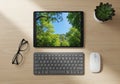 A workspace setup on a light wood desk features a tablet displaying a vivid image of green trees Royalty Free Stock Photo