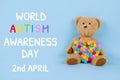 World Autism Awareness Day concept - sad teddy bear with multicolored heart sitting against blue background. Autism spectrum