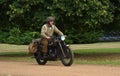 World War 2 despatch rider in uniform on classic motorcycle riding in park land.