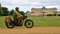 World War 2 despatch rider in uniform on classic motorcycle riding past stateley home.