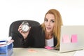 Worried attractive blond businesswoman holding alarm clock sitting at office desk working on laptop Royalty Free Stock Photo