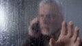 Worried elderly man looking through rainy window, talking on phone, waiting