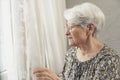 worried grey-haired caucasian grandma looking out of the window with white curtain Royalty Free Stock Photo