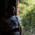 Worried middle-aged man with a glass of white wine in his hand illuminated by a dim evening light looking out a window of a rural Royalty Free Stock Photo