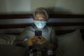 Worried middle aged woman 50s with grey hair and protective mask checking online news with mobile phone during covid-19 virus home Royalty Free Stock Photo