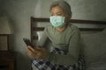 Worried middle aged woman 50s with grey hair and protective mask checking online news with mobile phone during covid-19 virus home Royalty Free Stock Photo