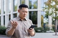 Worried senior asian male office worker businessman standing outside and using mobile phone. Disappointingly looks at Royalty Free Stock Photo