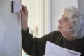 Worried Senior Woman With Bill Turning Down Central Heating Thermostat At Home In Energy Crisis Royalty Free Stock Photo