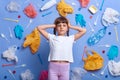 Worried upset little girl with braids wearing white t shirt posing against much plastic wastes on blue wall, standing hand on head Royalty Free Stock Photo