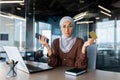 Worried and upset young muslim businesswoman in hijab holding phone and credit card. Sitting in the office at the desk Royalty Free Stock Photo