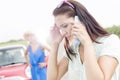 Worried woman using mobile phone while friend standing by broken down car on sunny day