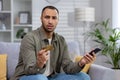 Worried young african american man sitting on couch at home and looking upset at camera, holding credit card and phone Royalty Free Stock Photo