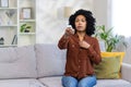 Worried young African American woman sitting on sofa at home and turning on air conditioner by remote control. He feels Royalty Free Stock Photo