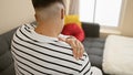 Worried young hispanic man sitting on sofa at home, suffering serious backache, a painful reality indoors Royalty Free Stock Photo