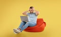 Worried young man with laptop on bean bag chair, yellow background