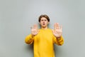 Worried young man in yellow clothes shows a palms gesture STOP and looks at the camera with a serious face on a gray wall Royalty Free Stock Photo