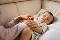 Worried young mother sitting on sofa beside her sick son with high fever. Mom measures temperature using thermometer of Royalty Free Stock Photo