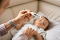 Worried young mother sitting on sofa beside her sick son with high fever. Mom measures temperature using thermometer of Royalty Free Stock Photo