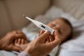 Worried young mother sitting on sofa beside her sick son with high fever. Mom measures temperature using thermometer of Royalty Free Stock Photo